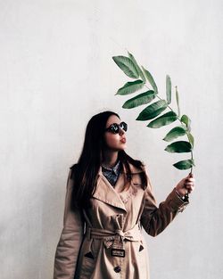 Woman holding twig with green leaves while standing against wall