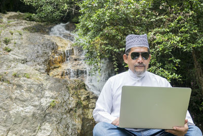 Mature man wearing sunglasses while using laptop against waterfall