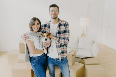 Portrait of couple with dog standing against wall in new house
