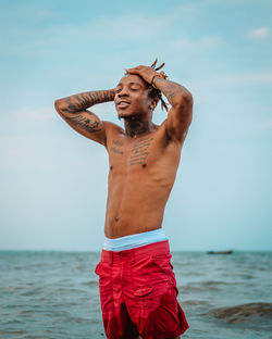 Portrait of shirtless man with tattoos standing at sea against sky