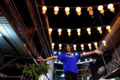 Low angle view of young woman standing against lighting equipments at night