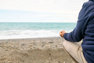 Rear view of man in casual clothes sitting in lotus pose on pebble winter beach and meditating