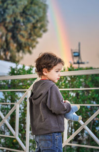 Cute boy standing by railing against rainbow