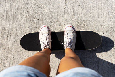 From above of crop anonymous woman in shorts and sneakers standing on skateboard on asphalt road during sunny day in summer