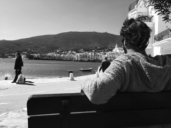 Rear view of man sitting on bench by river in city