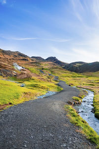 Pathway amidst grassy field by stream in volcanic valley against blue sky