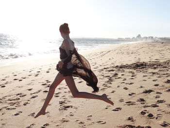 Full length side view of young woman jumping at beach on sunny day