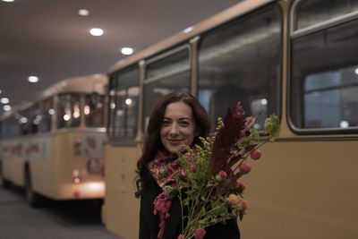 Portrait of woman holding bouquet while standing against bus on road at night