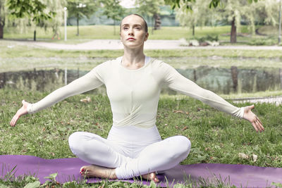 A young woman with long blonde hair does yoga in the summer in nature by the pond in the park.