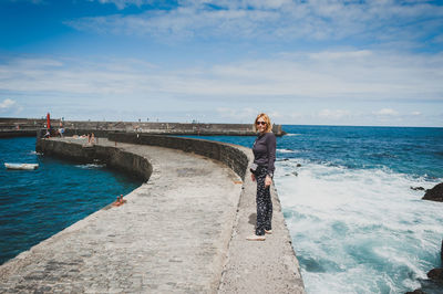 Full length portrait of woman standing on retaining wall by sea against sky