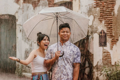 Smiling couple holding umbrella while standing against wall during rainfall