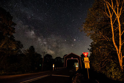 Sign board by road leading towards bridge against star field at night