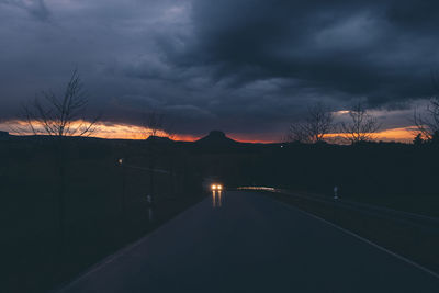 Illuminated road by silhouette mountain against sky at sunset