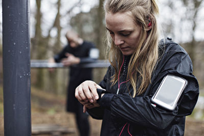 Female athlete checking smart watch at forest while man standing in background