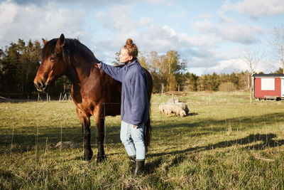 Side view of mid adult woman stroking horse while standing on grassy field against sky