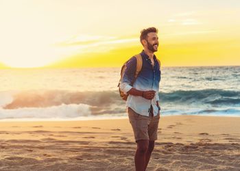 Rear view of man standing at beach against sky during sunset