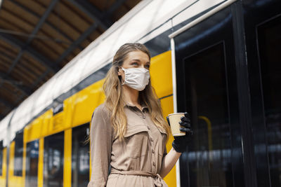 Woman wearing mask holding coffee while standing at railroad station