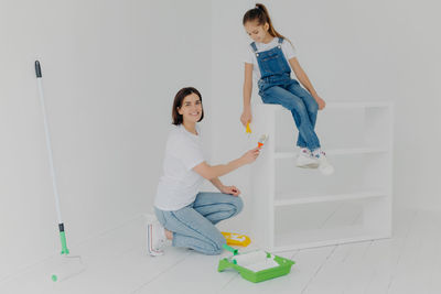 Full length of mother and daughter holding paint roller sitting at home