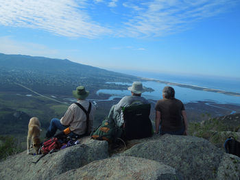 Rear view of people sitting on rocks by sea against sky