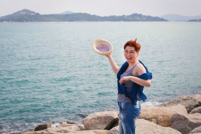 Portrait of smiling mature woman holding hat while standing on rock against sea at beach