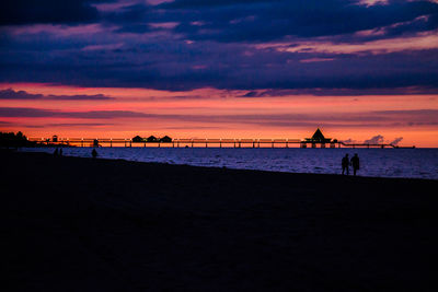 Silhouette pier on sea at beach against dramatic sky