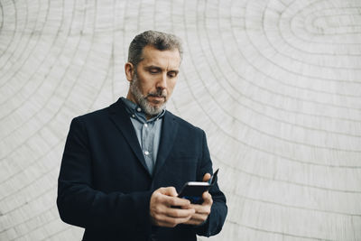 Mature businessman using smart phone against wall at office