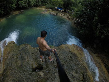 Rear view of man with monopod standing on cliff by river in forest