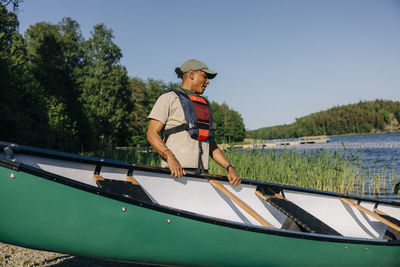 Male counselor wearing life jacket holding kayak near lake at summer camp