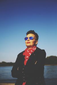 Smiling woman wearing sunglasses while standing by lake against clear blue sky