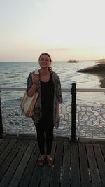 Portrait of smiling young woman with ice cream standing on pier over beach