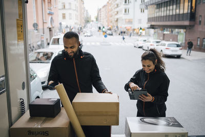 Smiling coworkers using technologies while examining packages in delivery van