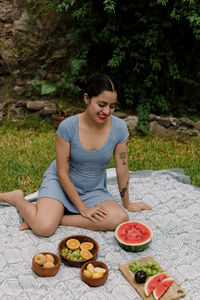 Full length of woman with fruits sitting on picnic blanket in yard