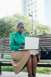 Concentrated young african american female freelancer with short dyed hair in stylish clothes sitting on wooden bench in park and working remotely on laptop