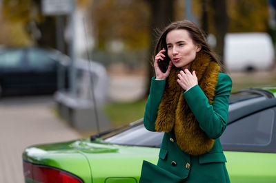 A woman talking on the phone while standing by a green car parked on a city street in the background