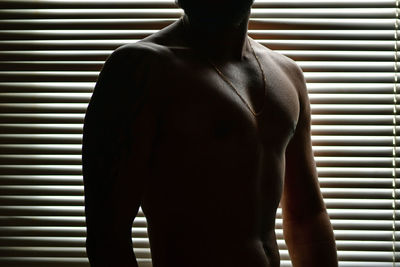 Shirtless man standing against window blinds