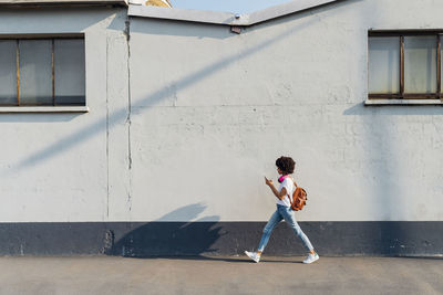 Young woman using smart phone walking by wall on footpath