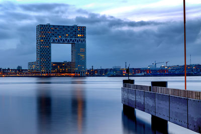 Illuminated city at waterfront against cloudy sky