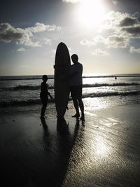 Silhouette father holding surfboard while standing by son against sea during sunset