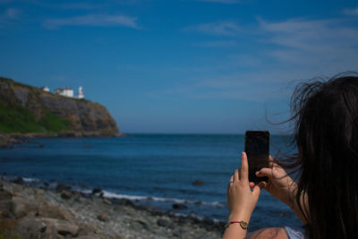 Rear view of woman photographing sea with smart phone against sky