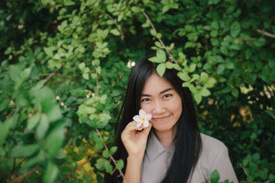 Portrait of smiling young woman holding flower by tree