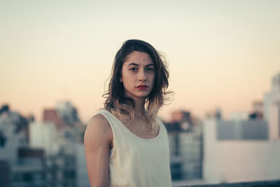 Portrait of young woman standing against clear sky in city during sunset