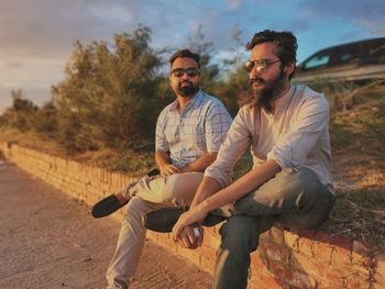 Portrait of two mid adult men sitting and hanging out together on a small wall during sunset