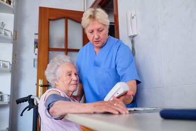 Caring nurse with professional tonometer measuring blood pressure to aged woman sitting at table with food in kitchen at home