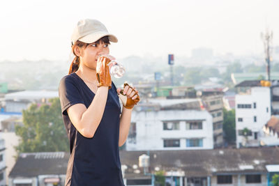 Young woman drinking water while standing by cityscape against clear sky