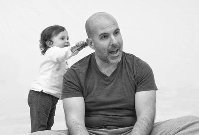 Cute girl combing hair of father while sitting against wall at home