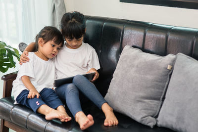 Full length of cute sisters using digital tablet while sitting on sofa at home