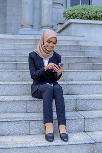 Full length of a young woman using phone while sitting on staircase