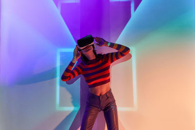 Woman in modern black vr goggles standing with raised arms in futuristic studio with glowing lights while exploring virtual reality
