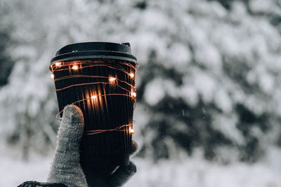 Female hand in winter warm gloves holding black eco paper cup with garland shiny light. creative 