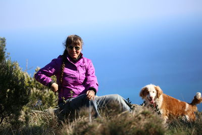 Portrait of woman sitting with dog on grassy field against blue sky